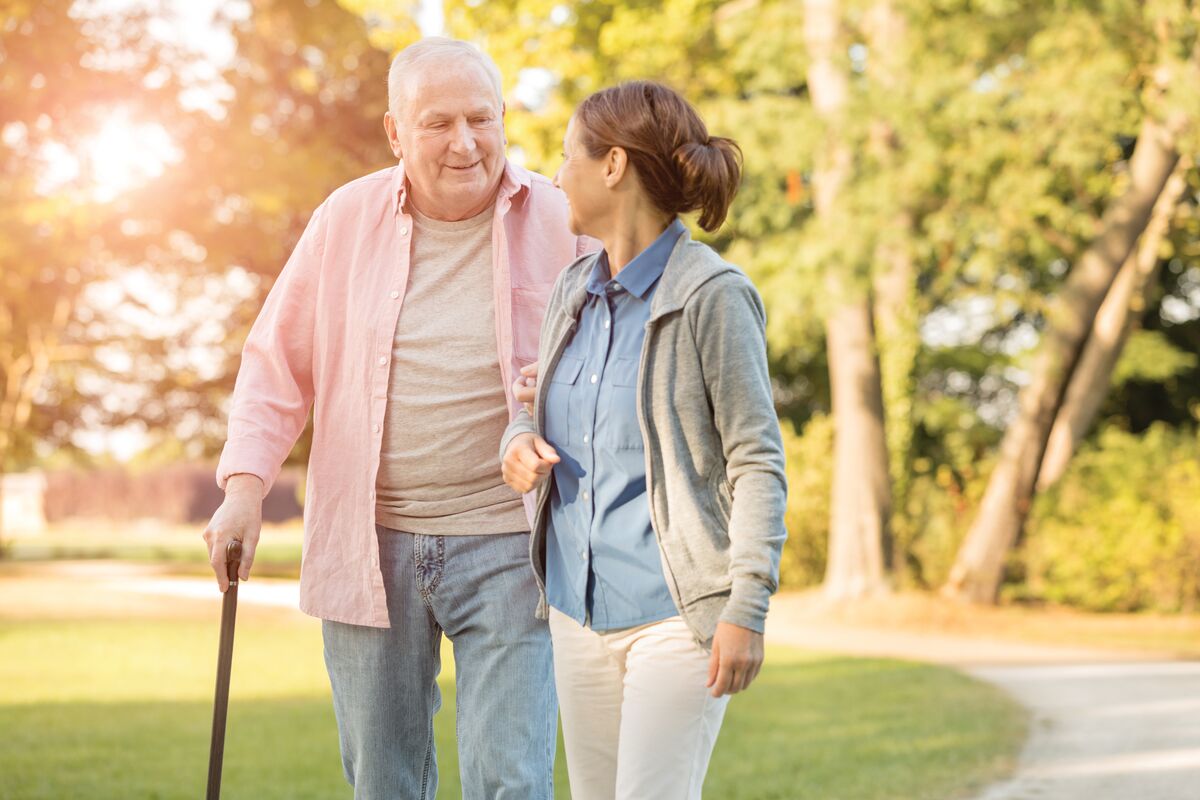 Elderly man with a cane walking outdoors with a woman supporting him in a park.