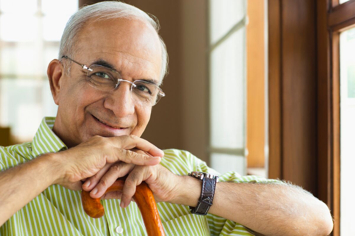 Older man wearing glasses, leaning on a wooden cane, and smiling indoors near a window.