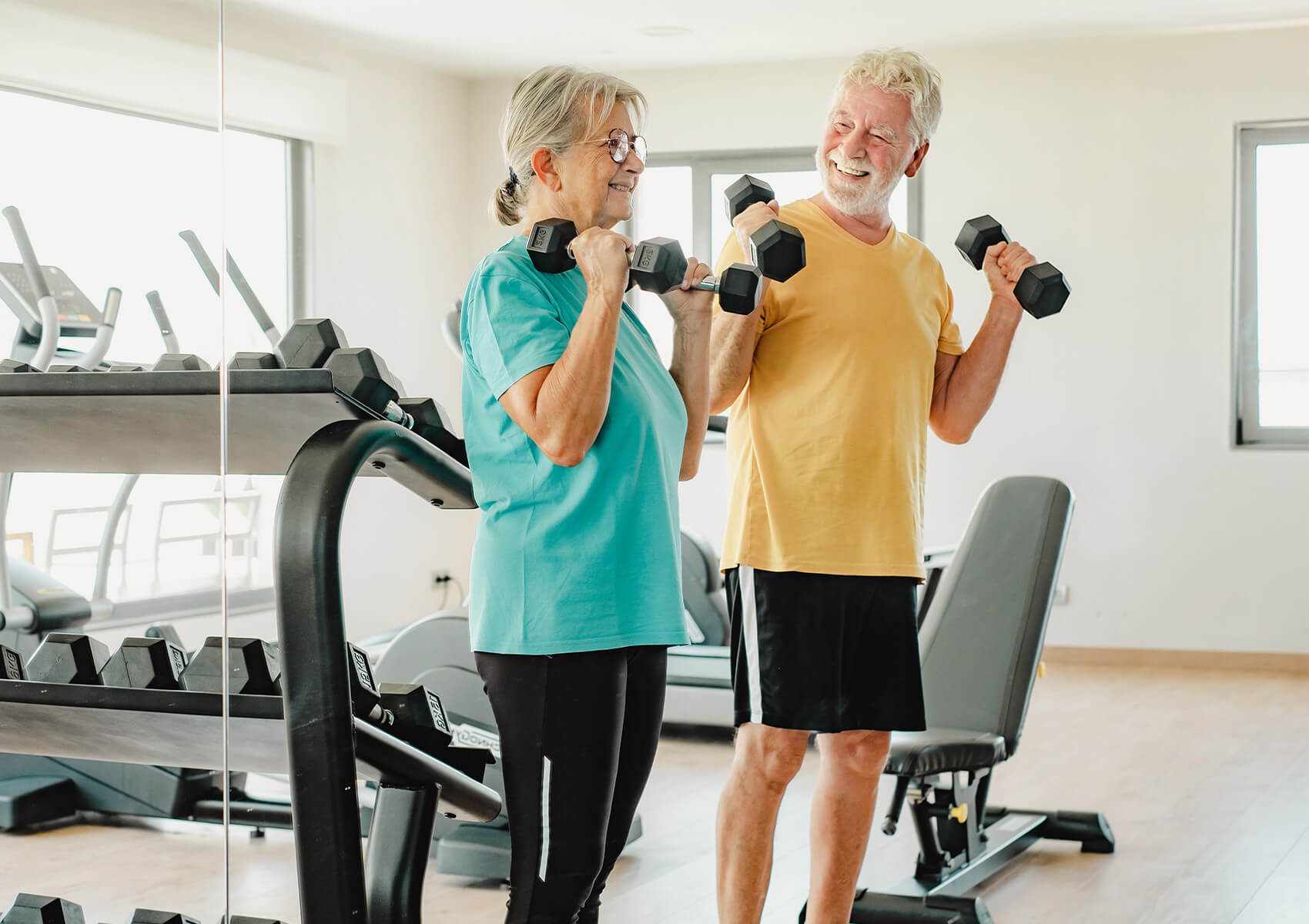 Smiling senior couple lifting dumbbells in a bright, modern fitness center.
