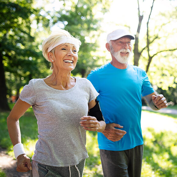 Smiling seniors jogging in a lush green park on a sunny day.