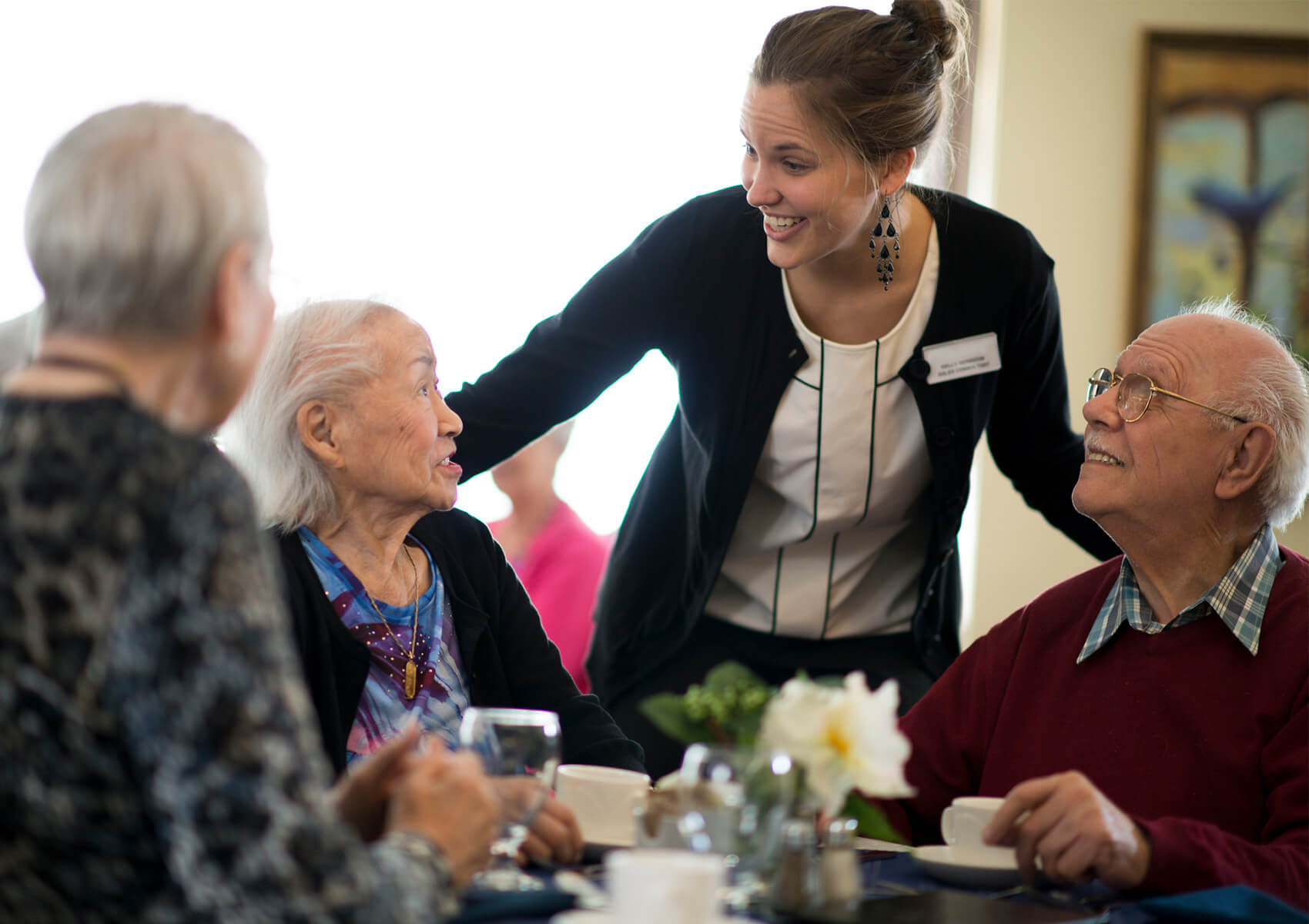 Smiling caregiver engages with three seniors around a table in a communal hall.