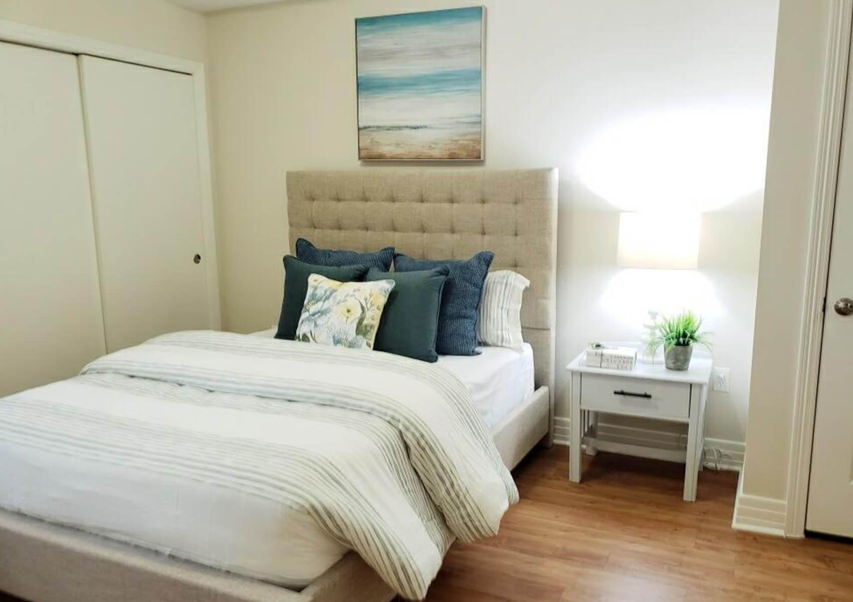 Cozy living unit bedroom with a bed, nightstand, lamp, and calming beach artwork.