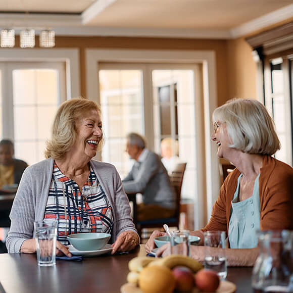 Two senior women sharing a meal and laughing in a dining area of a community building.