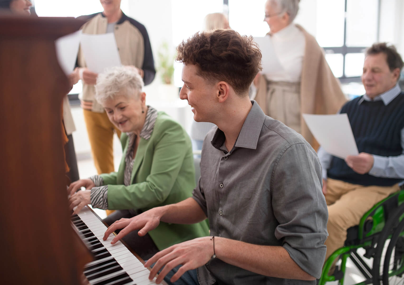 Young man plays piano with elderly woman in green jacket in a communal room.