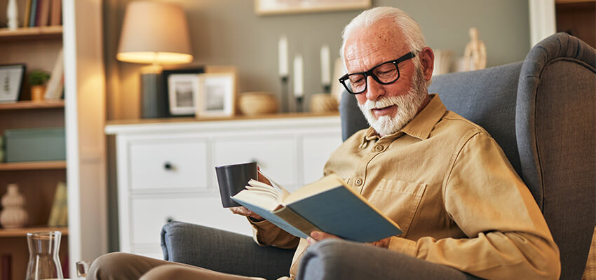 Elderly man reading a book in a cozy living room, holding a coffee mug.