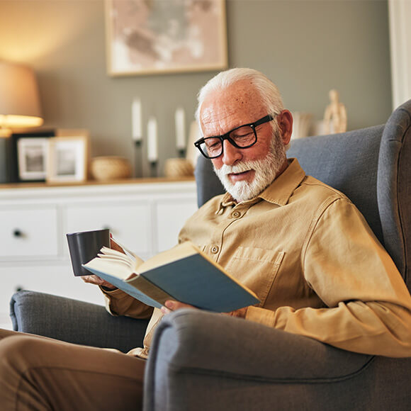 Senior man in glasses reading a book and holding a mug, sitting in a cozy armchair.