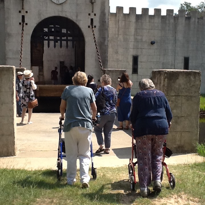 Visitors, including elderly individuals with walkers, enter a castle-like building through a drawbridge.