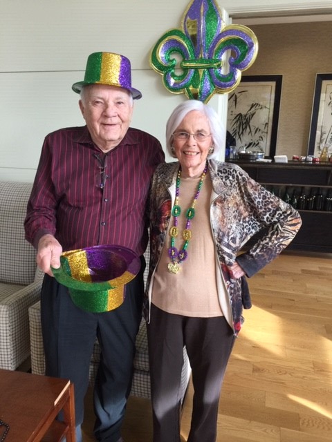 Two older adults dressed festively with a Mardi Gras theme, including colorful hats and beads.