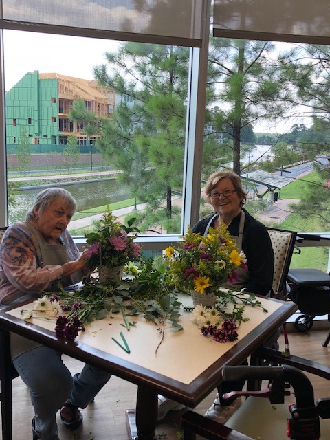 Two elderly women arrange colorful flowers at a table with a nature view in the background.