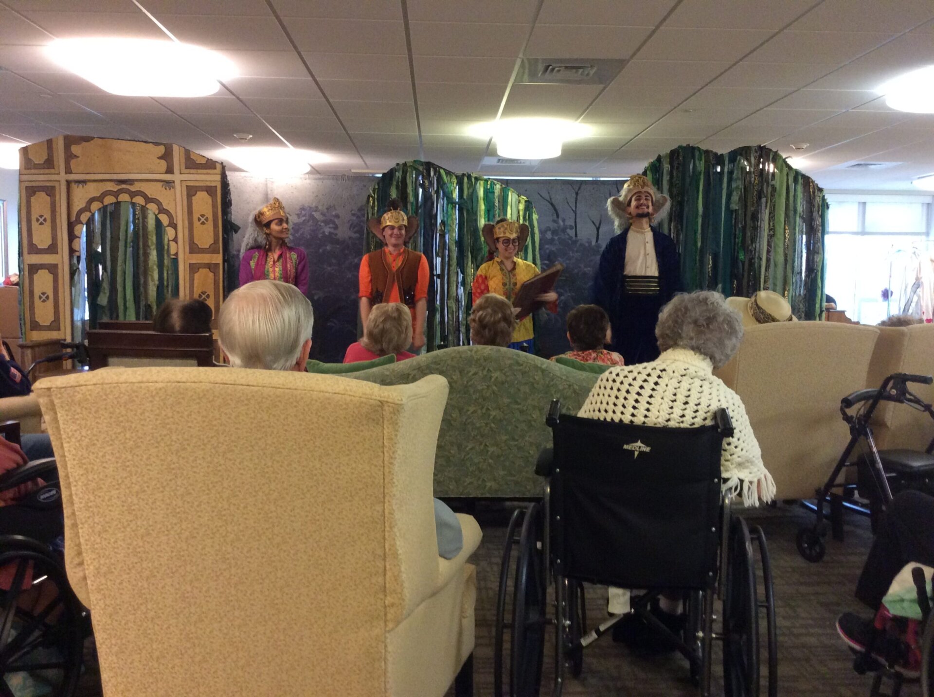 Performers in colorful costumes entertain residents in a common area with vibrant decorations.