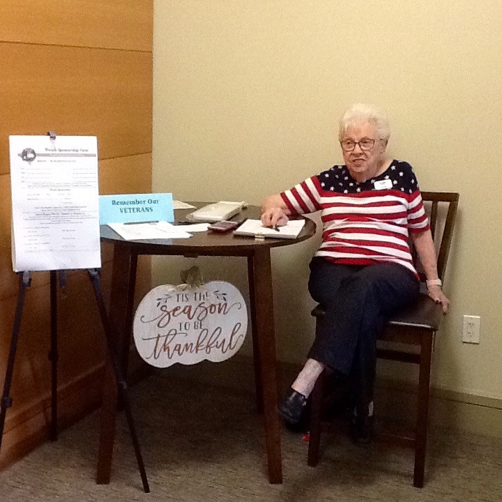 Elderly woman sitting at a table with patriotic decor and a sign reading Tis the Season to be Thankful.