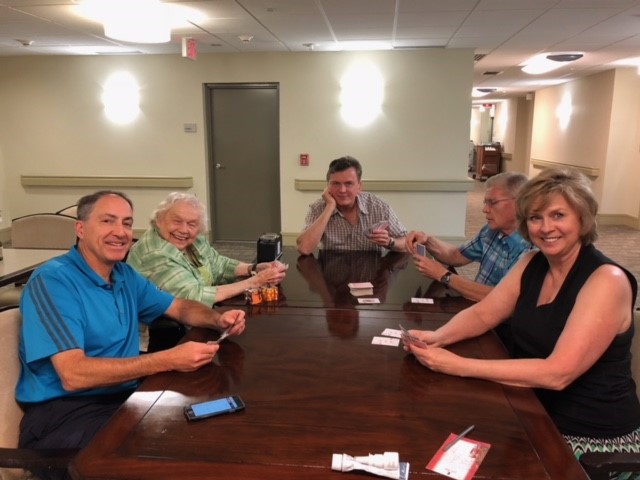 Five people sitting around a wooden table playing cards in a well-lit communal area.