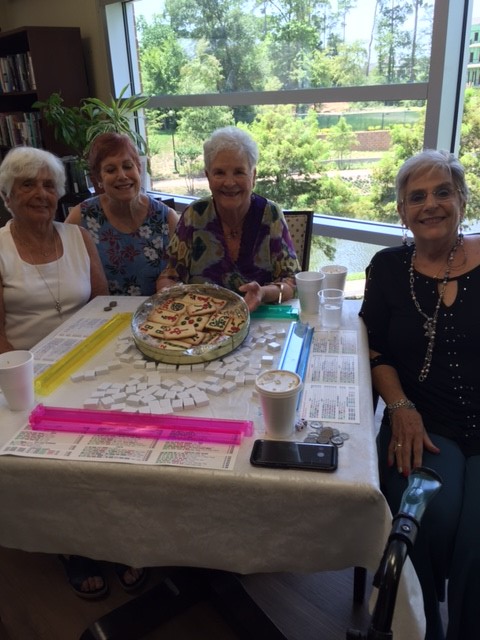 Four women smiling while playing mahjong in a large windowed room with greenery outside.