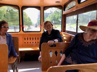 Three women sitting inside a wooden trolley with windows and greenery visible outside.