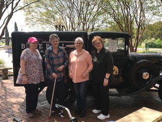 Four women standing in front of a vintage truck on a brick patio with trees in the background.