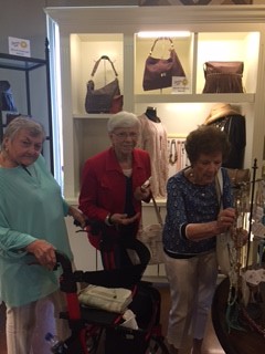 Three older women shopping for handbags inside a well-lit store, one using a walker.