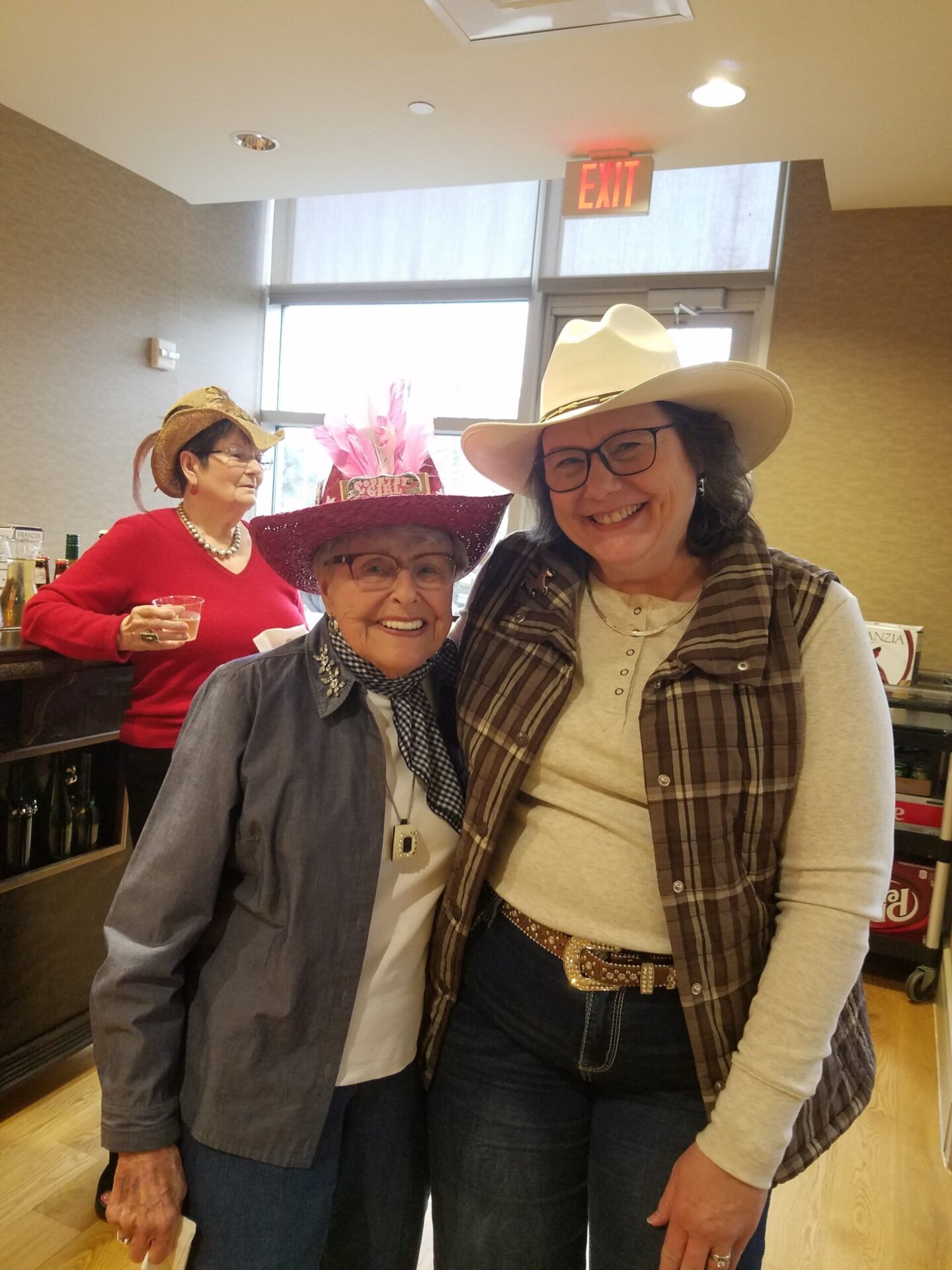 Two women wearing festive hats smiling indoors with another person in the background.